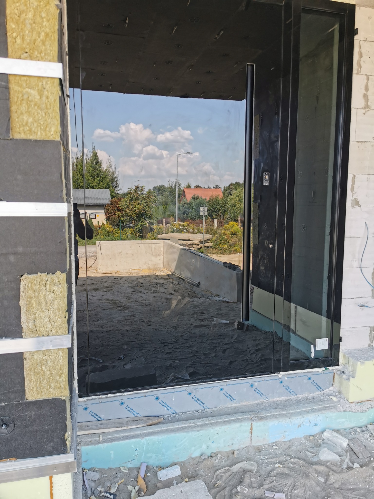 Montaż drzwi przesuwnych z czarną ramą w surowym stanie budynku, widoczne ocieplenie ścian i piasek na zewnątrz.