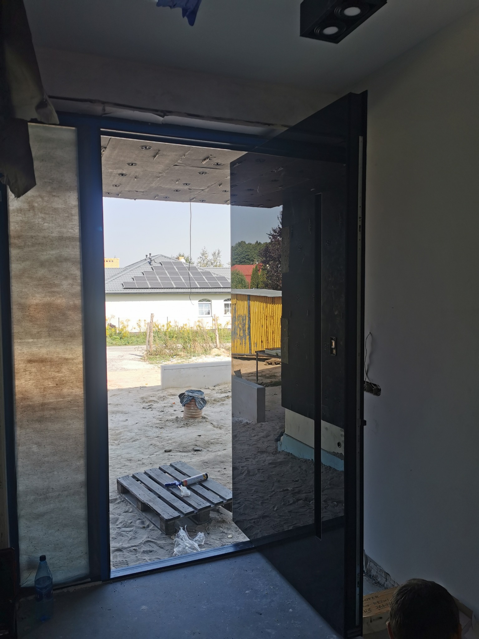 Montaż czarnej ramy drzwiowej z dużym przeszkleniem w surowym wnętrzu; widok na zewnątrz z paletą narzędzi i dom z panelami słonecznymi w tle.