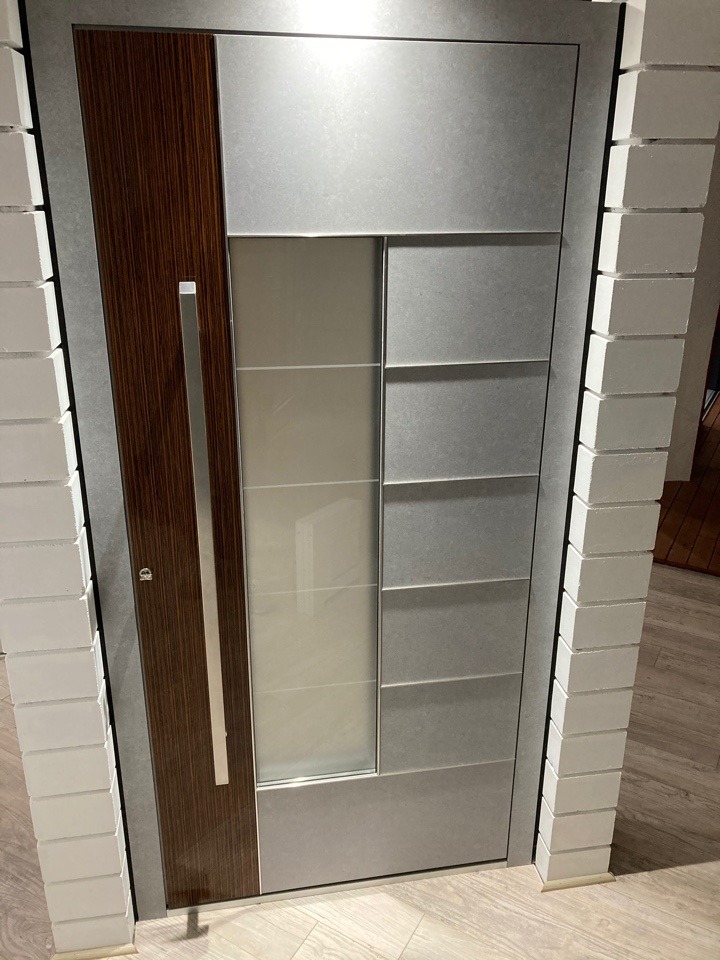 Nowoczesne drzwi wejściowe z panelem drewnopodobnym i szybą mleczną, osadzone w szarej ramie, widoczne w ekspozycji na tle białych cegiełek.