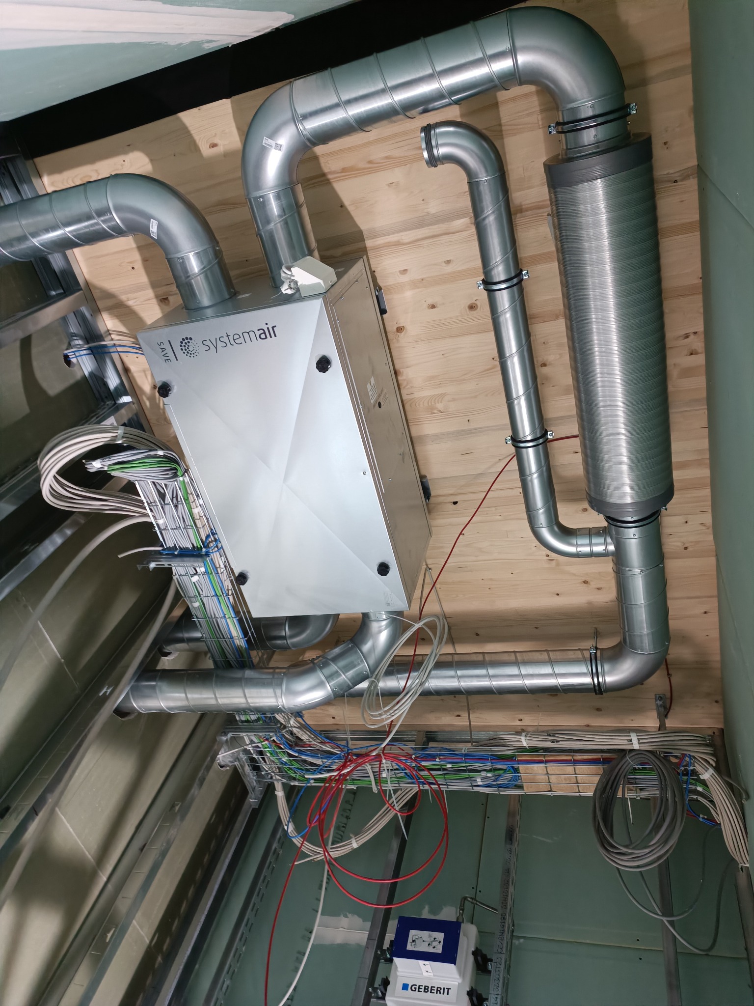 Systemair centrala wentylacyjna z podłączonymi przewodami wentylacyjnymi i elektrycznymi, zamontowana pod drewnianym stropem, widok instalacji w trakcie montażu.