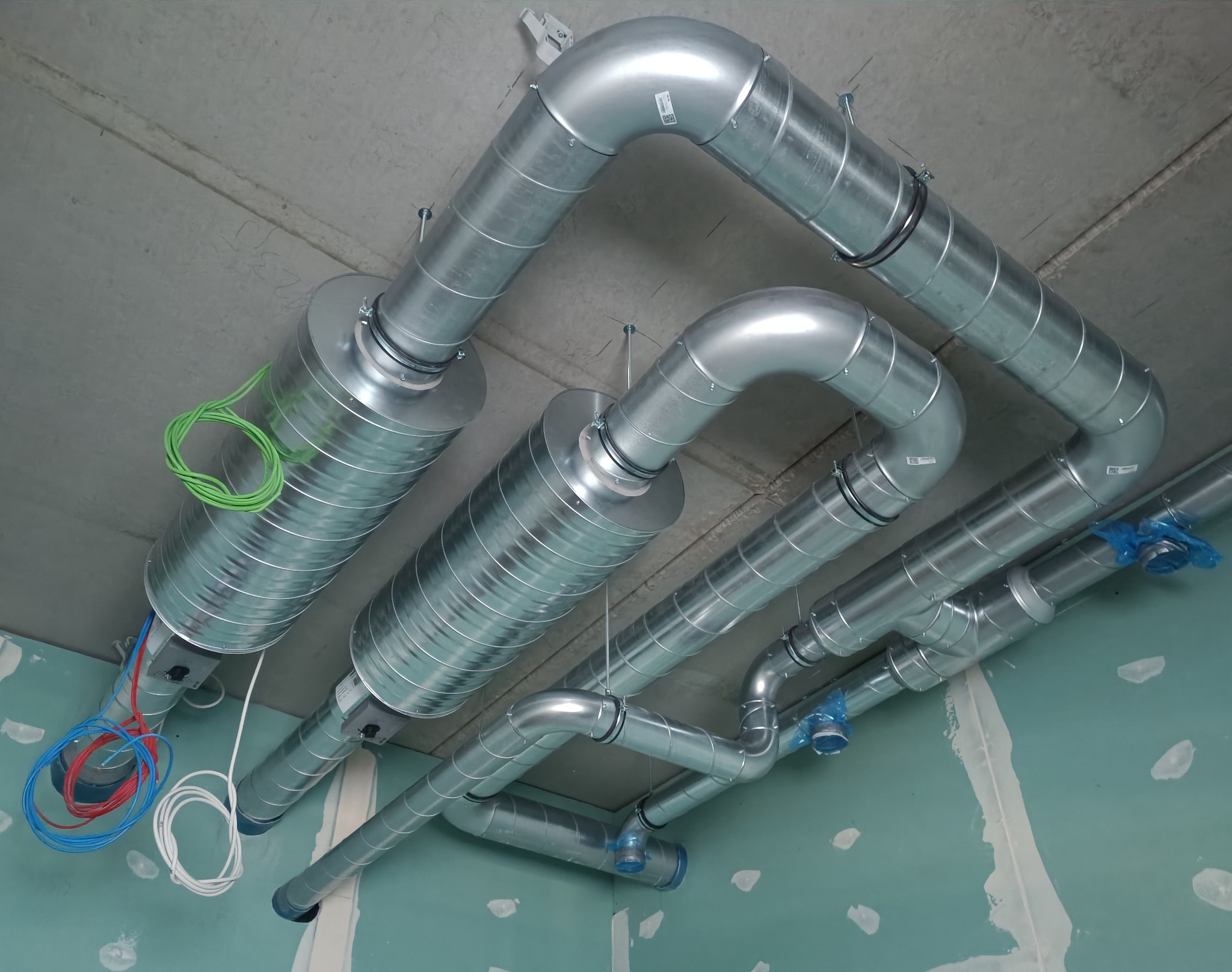 Widok z dołu na system srebrnych rur wentylacyjnych z przewodami elektrycznymi w trakcie montażu w surowym wnętrzu z płyt gipsowo-kartonowych.