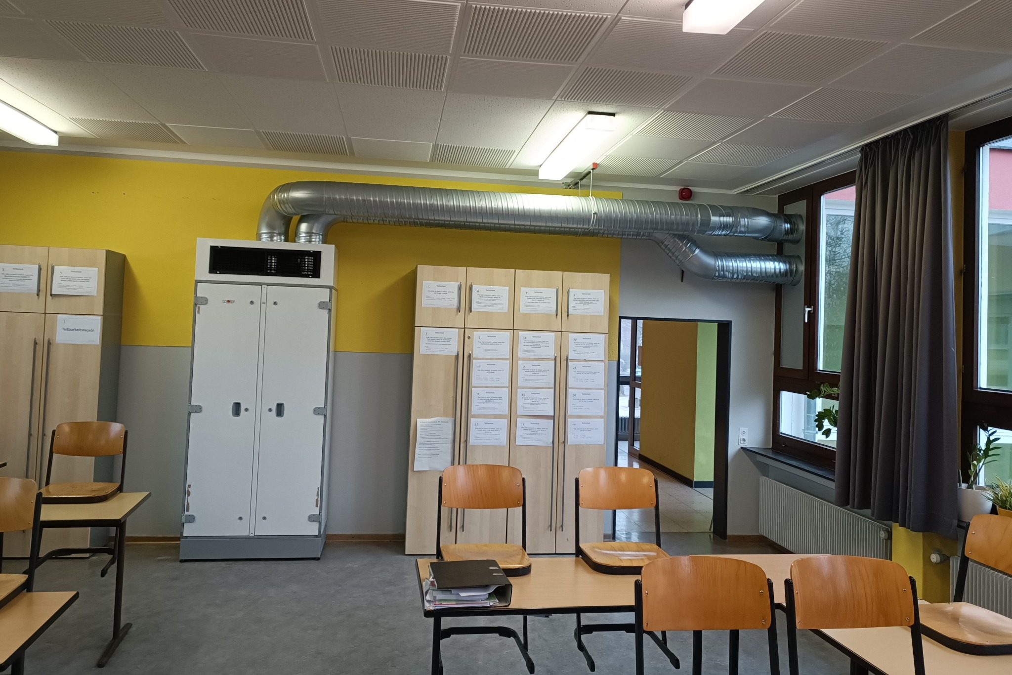 Wnętrze klasy z widocznym systemem kanałów wentylacyjnych pod sufitem, połączonych z białą szafą sterowniczą przy żółtej ścianie, oraz meblami szkolnymi.