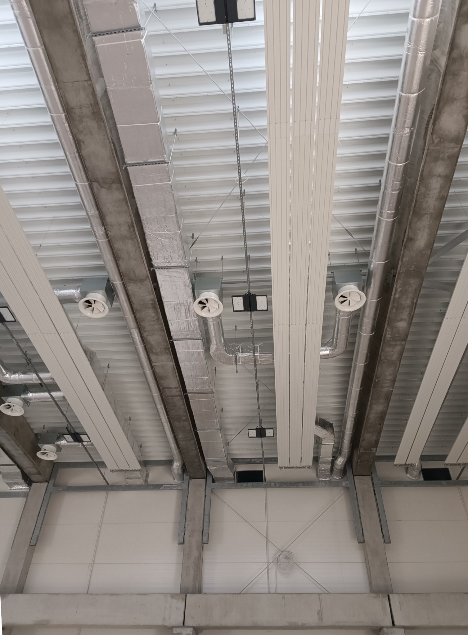 Widok z dołu na sufit hali przemysłowej z zamontowanymi kanałami wentylacyjnymi, izolowanymi rurami, anemostatami i oświetleniem LED.