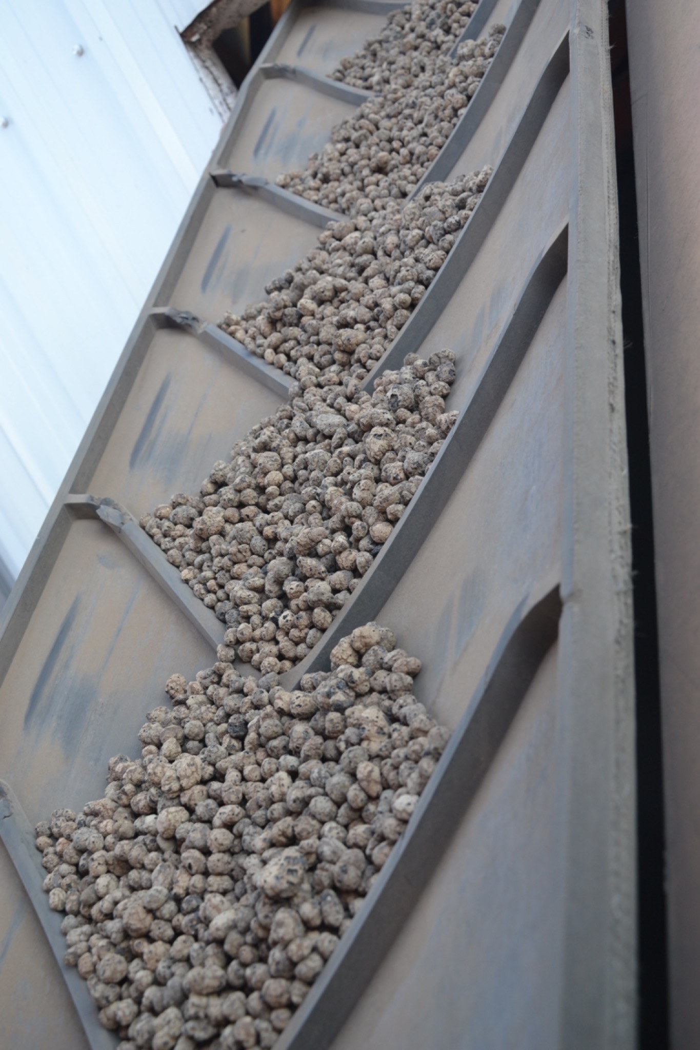 Powiększony widok na szary, metalowy podajnik wypełniony szarobrązowymi, okrągłymi granulkami perlitu.