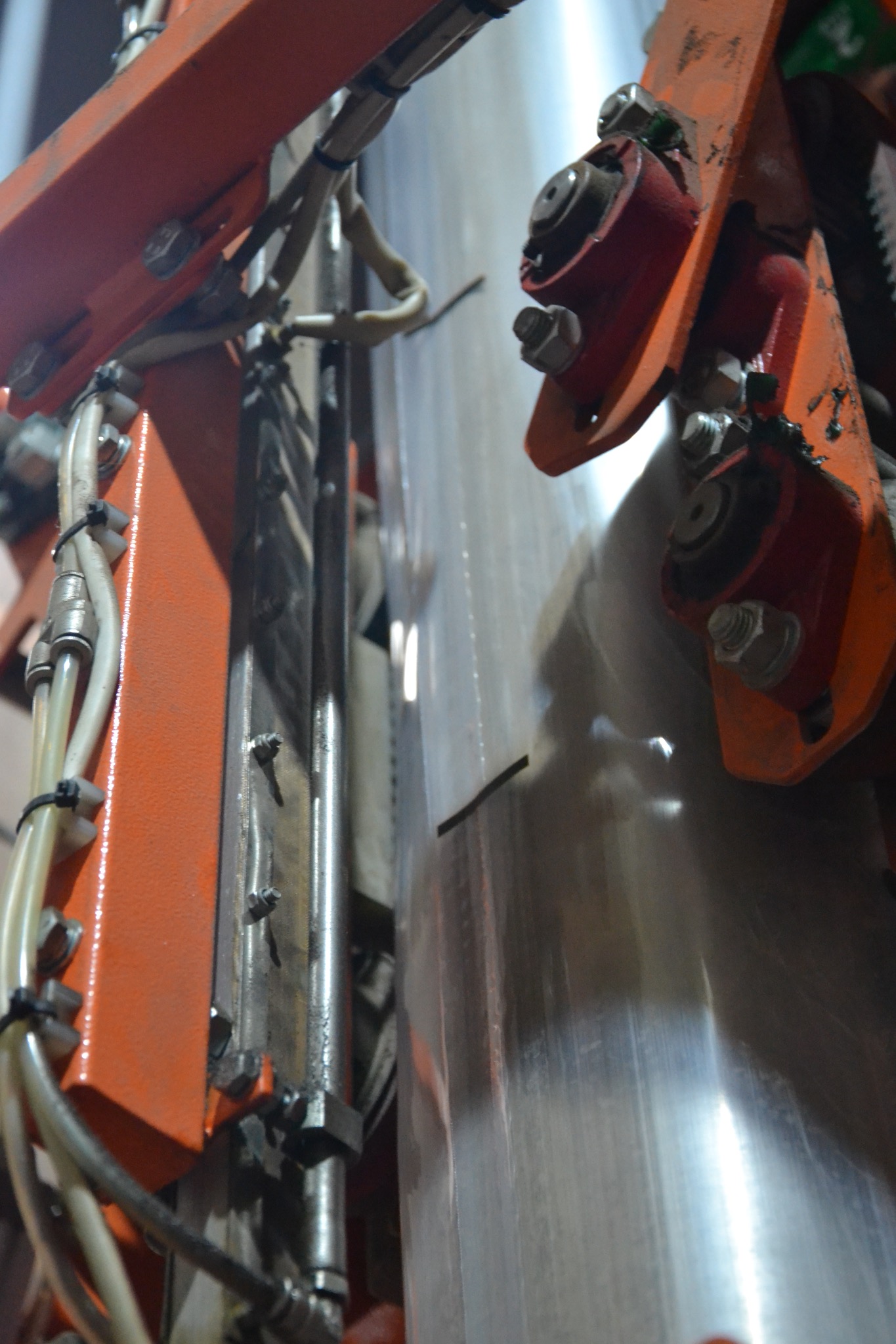 Fragment maszyny do pakowania z pomarańczowymi elementami konstrukcyjnymi, widoczne przewody i metalowe części, w tle przezroczysta folia.