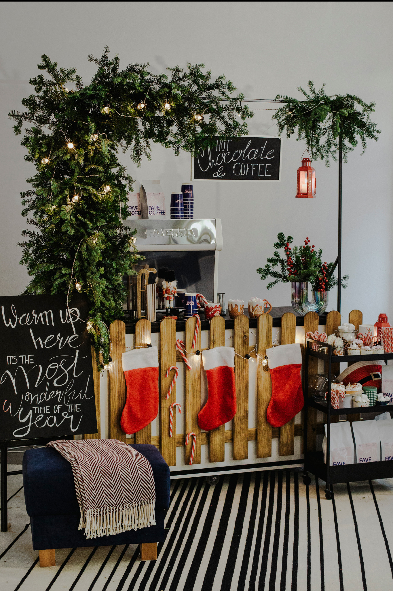 Świąteczny bufet z napojami: dekoracje z gałązek świerkowych z lampkami, tablica z napisem 'Hot Chocolate & Coffee', czerwone skarpety zawieszone na płotku, laski cukrowe, ciasteczka i muffinki...