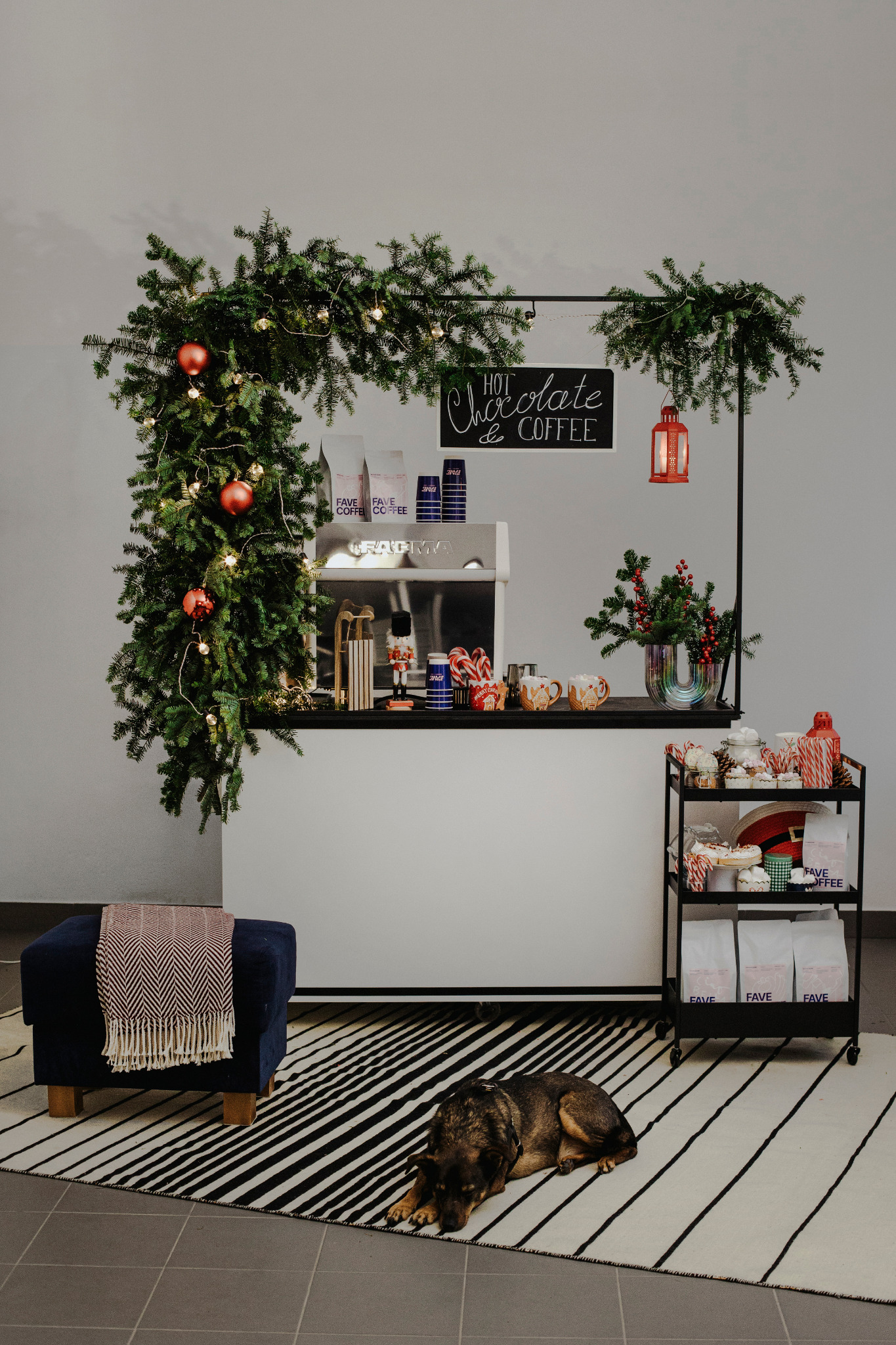 Świątecznie udekorowany bar kawowy z ekspressem, słodyczami i dekoracjami bożonarodzeniowymi, na podłodze leży pies rasy owczarek niemiecki na dywanie w czarno-białe pasy, obok pufa z pledem.