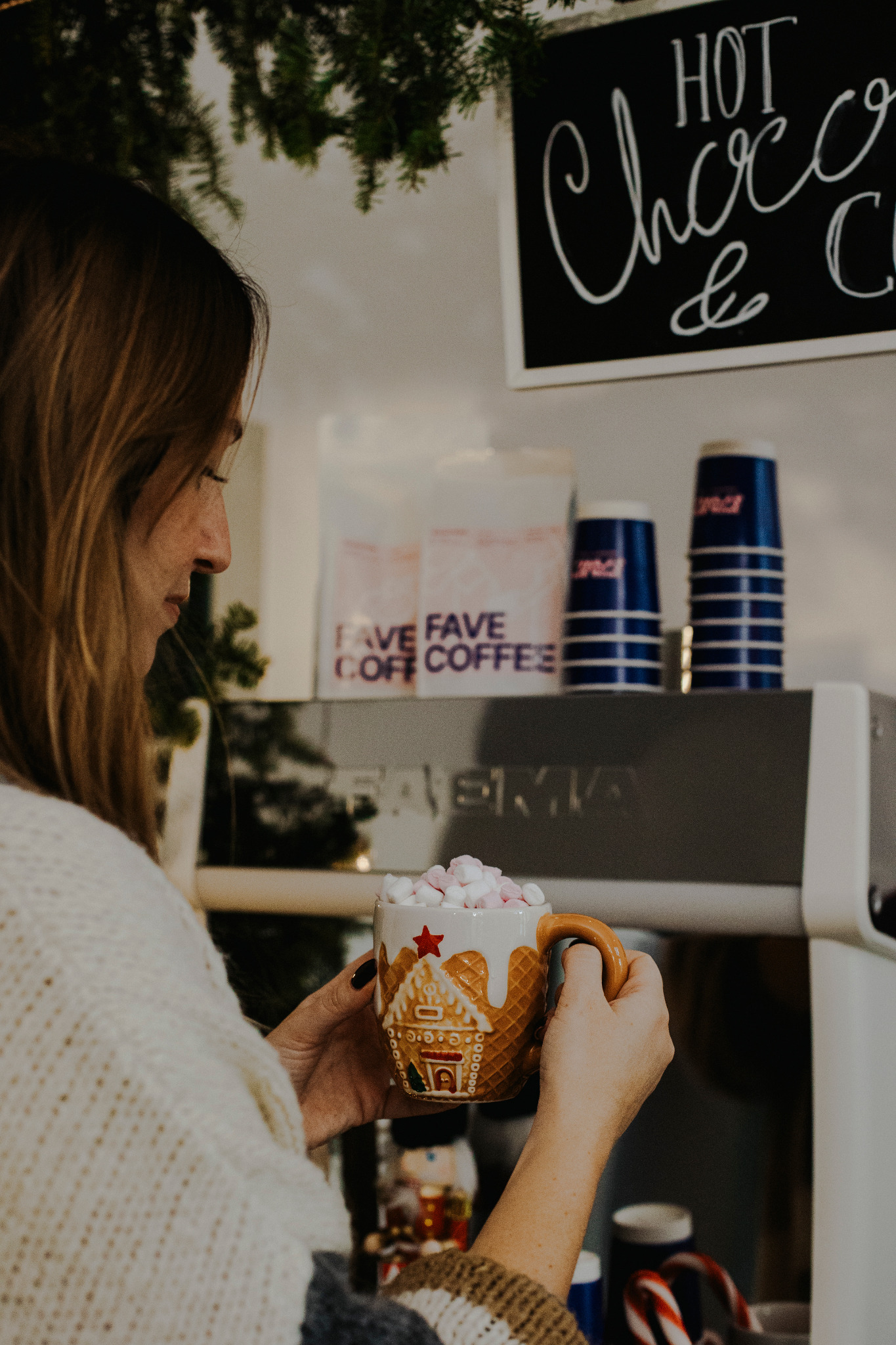 Kobieta trzyma świąteczny kubek z piernikowym domkiem, wypełniony piankami, na tle tablicy z napisem 'Hot Choco & C', obok ekspresu do kawy i opakowań kawy.