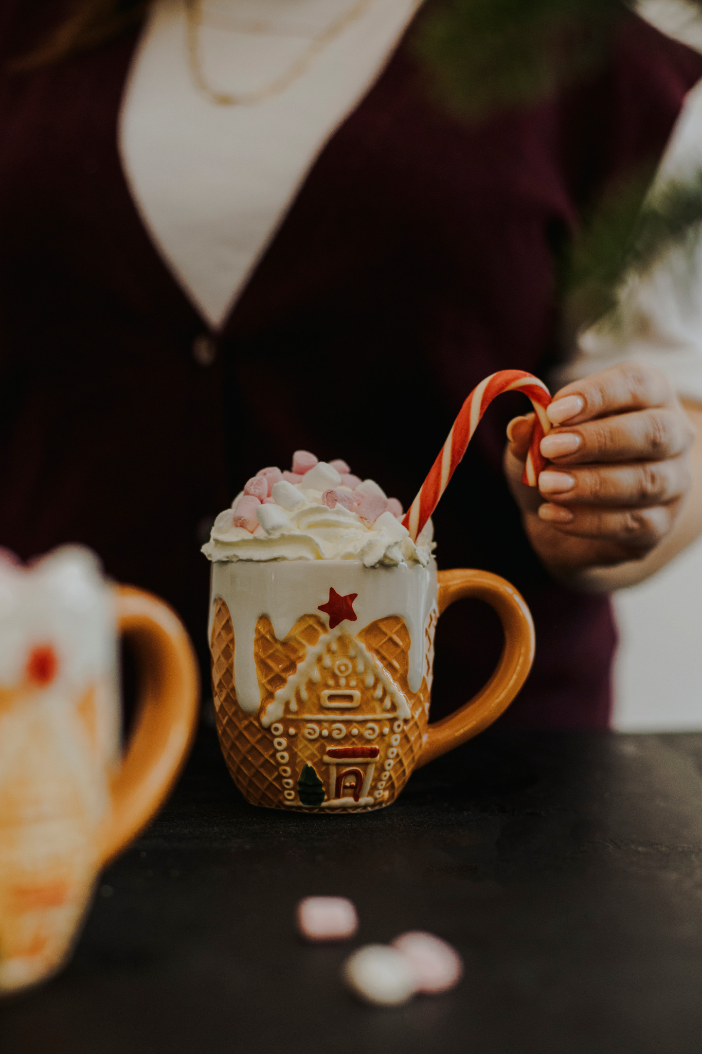 Kawa w ceramicznym kubku w kształcie piernikowego domku, udekorowana bitą śmietaną, piankami i laską cukrową, trzymana w dłoni.