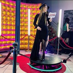 Magic Party Bolesławiec Dmuchańce Namioty Dawid Kłos - Kobieta w czarnej sukience i kapeluszu pozuje z rekwizytem 'Gruby Melanż' na platformie obrotowej z neonowym oświetleniem, tło z błyszczących paneli i lamp LED.