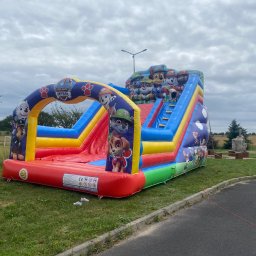 Magic Party Bolesławiec Dmuchańce Namioty Dawid Kłos - Kolorowy, dmuchany zamek ze zjeżdżalnią i motywami z bajki Psi Patrol, ustawiony na trawie obok asfaltowej drogi w pochmurny dzień.