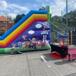 Magic Party Bolesławiec Dmuchańce Namioty Dawid Kłos - Kolorowa dmuchana zjeżdżalnia z postaciami z bajki Psi Patrol, obok stół z maszyną do popcornu i wata cukrowa na tle budynku i ogrodzenia.