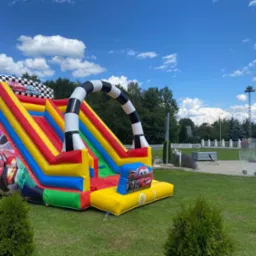 Kolorowy dmuchany zamek ze zjeżdżalnią, motyw Cars, ustawiony na trawniku w słoneczny dzień, z widocznym niebem i drzewami w tle.