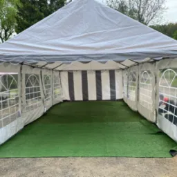 Rozłożony biało-szary namiot z przezroczystymi oknami i sztuczną zieloną trawą wewnątrz, widok z zewnątrz na żwirowe podłoże.