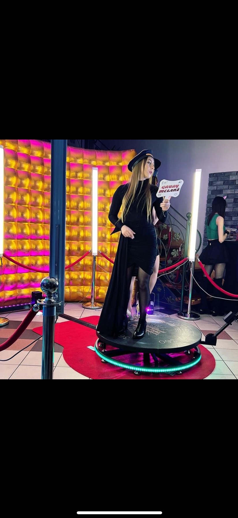 Kobieta w czarnej sukience i kapeluszu pozuje z rekwizytem 'Gruby Melanż' na platformie obrotowej z neonowym oświetleniem, tło z błyszczących paneli i lamp LED.