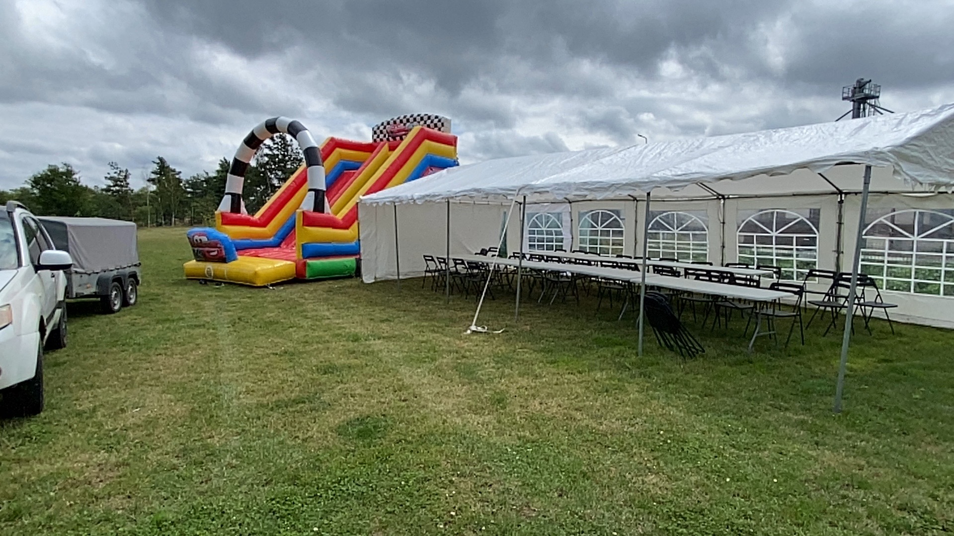 Rozłożony biały namiot z oknami i ustawionymi stołami oraz krzesłami na trawie, obok kolorowa dmuchana zjeżdżalnia z motywem wyścigowym, w tle samochód z przyczepą i pochmurne niebo.