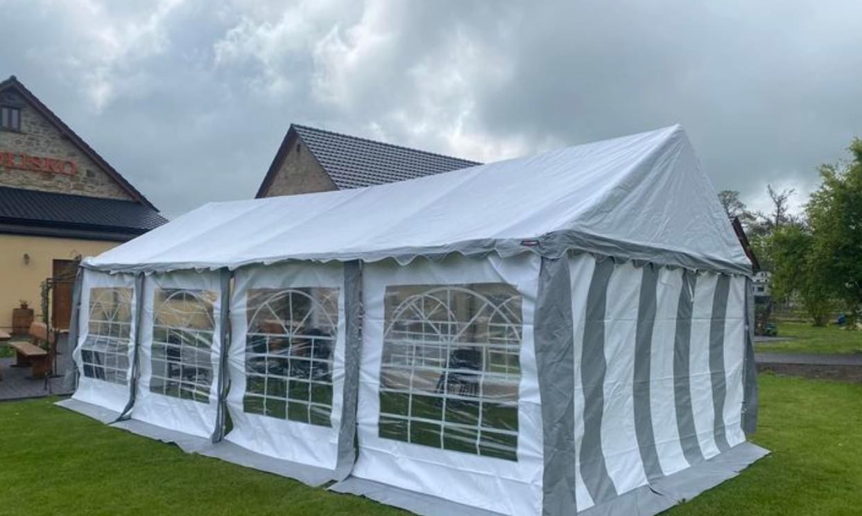 Rozłożony biały namiot z szarymi pasami na trawniku przed budynkiem z cegły pod pochmurnym niebem.