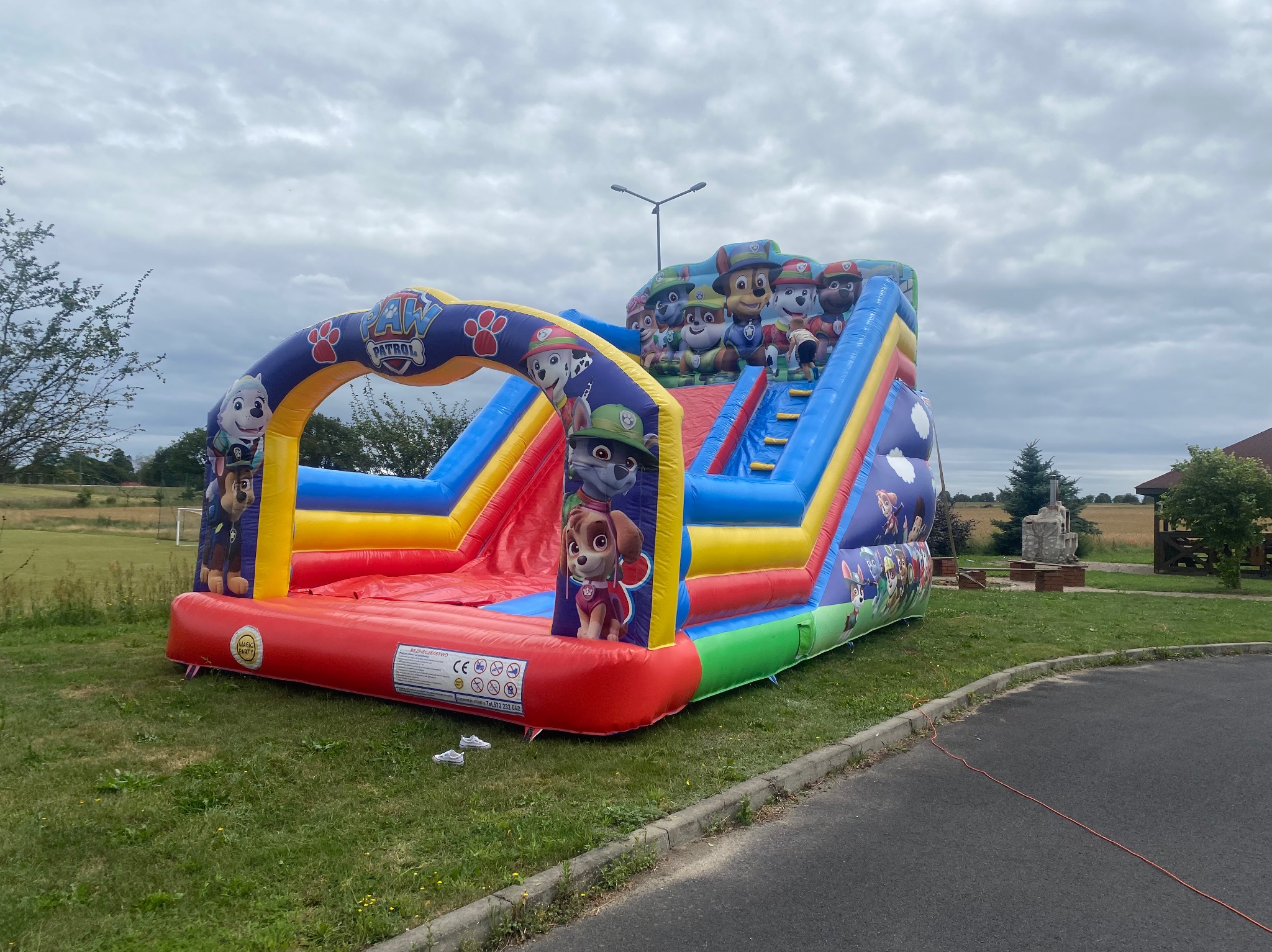 Kolorowy, dmuchany zamek ze zjeżdżalnią i motywami z bajki Psi Patrol, ustawiony na trawie obok asfaltowej drogi w pochmurny dzień.