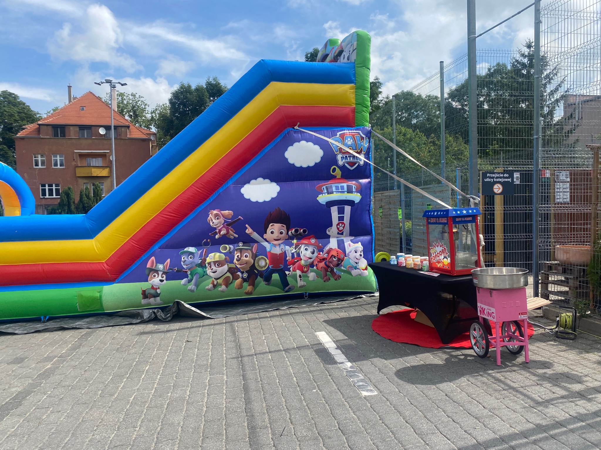 Kolorowa dmuchana zjeżdżalnia z postaciami z bajki Psi Patrol, obok stół z maszyną do popcornu i wata cukrowa na tle budynku i ogrodzenia.