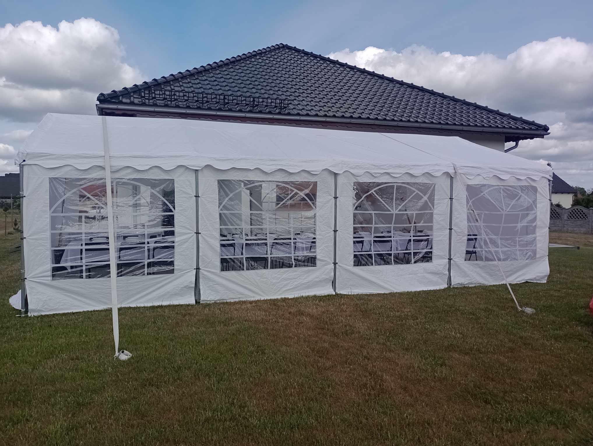 Biały namiot imprezowy z przezroczystymi oknami ustawiony na trawniku przed domem z ciemną dachówką. Wewnątrz widoczne stoły i krzesła.