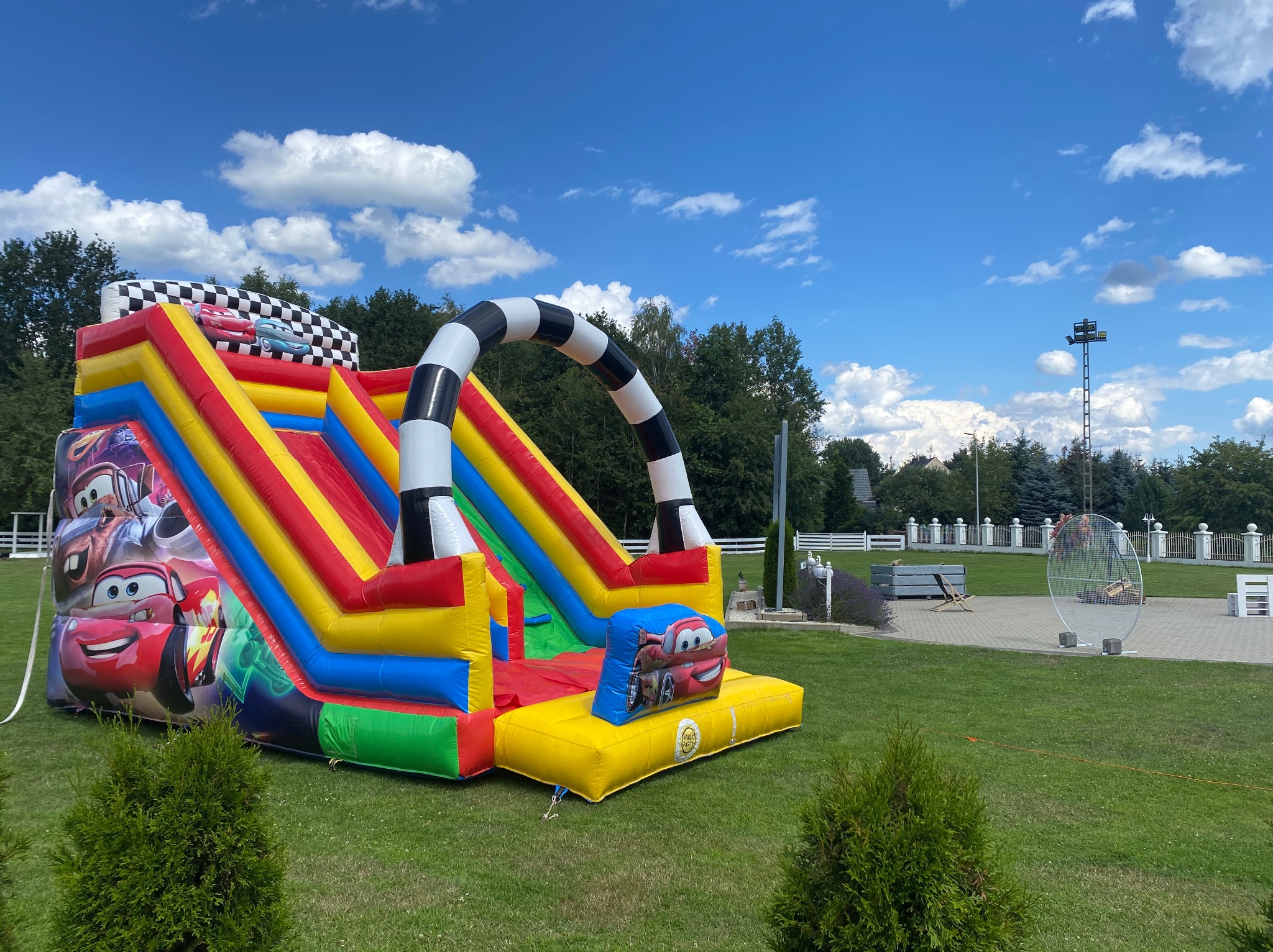 Kolorowy dmuchany zamek ze zjeżdżalnią, motyw Cars, ustawiony na trawniku w słoneczny dzień, z widocznym niebem i drzewami w tle.