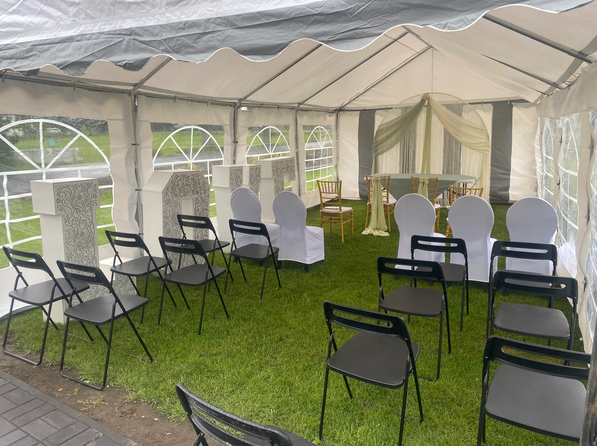 Ustawione czarne krzesła i białe dekoracje wewnątrz białego namiotu z przezroczystymi oknami, zielona trawa jako podłoże, okrągły stół z zielonym obrusem i złotymi krzesłami w tle.