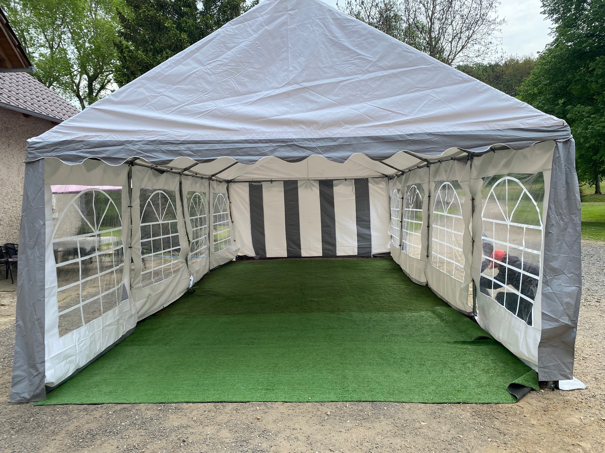 Rozłożony biało-szary namiot z przezroczystymi oknami i sztuczną zieloną trawą wewnątrz, widok z zewnątrz na żwirowe podłoże.