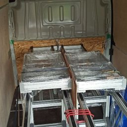 JOB COMPANY Sp. Z O. O. - Transport aluminiowych konstrukcji pod panele fotowoltaiczne w przestrzeni ładunkowej busa, zabezpieczone folią i spięte czerwoną gumą.