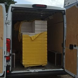 JOB COMPANY Sp. Z O. O. - Transport białych paneli fotowoltaicznych w żółtej folii ochronnej, załadowanych do białego busa z otwartymi drzwiami, widok z tyłu.