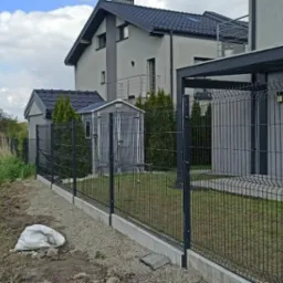 Czarny panelowy system ogrodzeniowy z podmurówką betonową, oddzielający posesję z domem w nowoczesnym stylu od pola uprawnego, z workiem gruzu przy fundamencie.