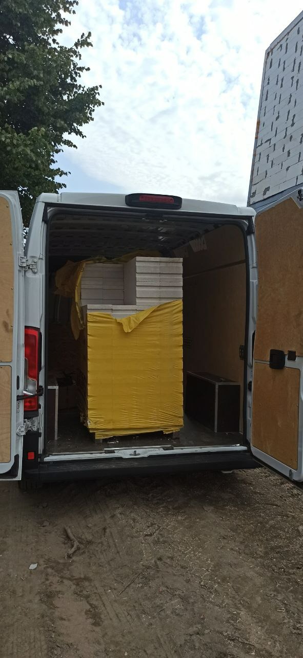 Transport białych paneli fotowoltaicznych w żółtej folii ochronnej, załadowanych do białego busa z otwartymi drzwiami, widok z tyłu.