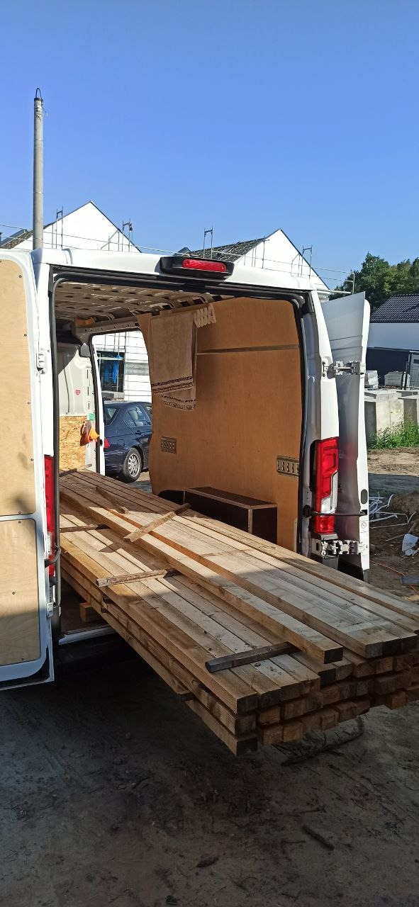 Otwarty bagażnik białego busa z widocznym stosem drewnianych belek na wysuwanej platformie, przygotowanych do montażu ogrodzenia na budowie w tle.
