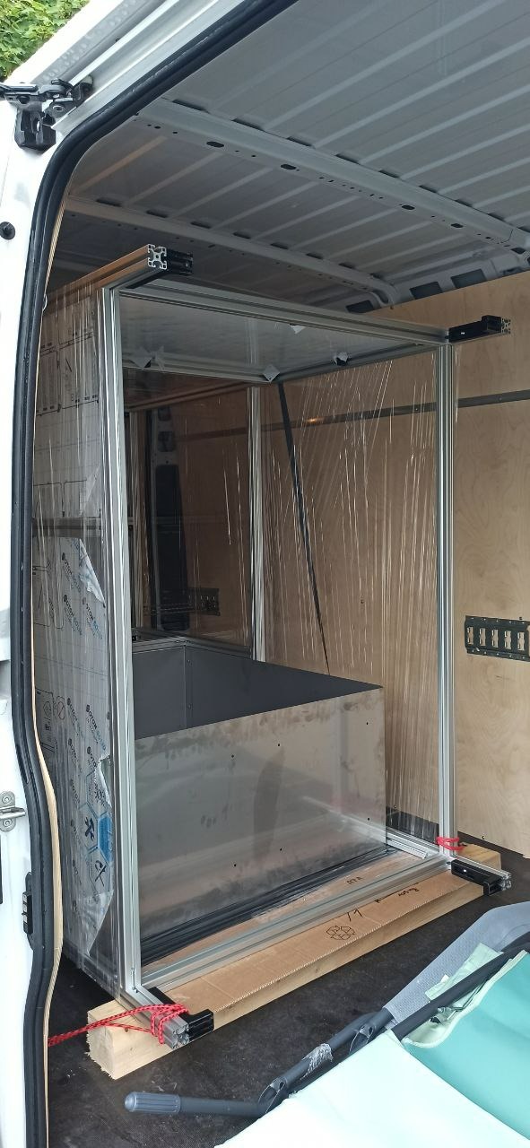 Wnętrze furgonetki z zamontowaną aluminiową konstrukcją, prawdopodobnie częścią większego urządzenia, zabezpieczoną folią i przymocowaną do podłogi za pomocą drewnianych klocków i czerwonego sznurka.