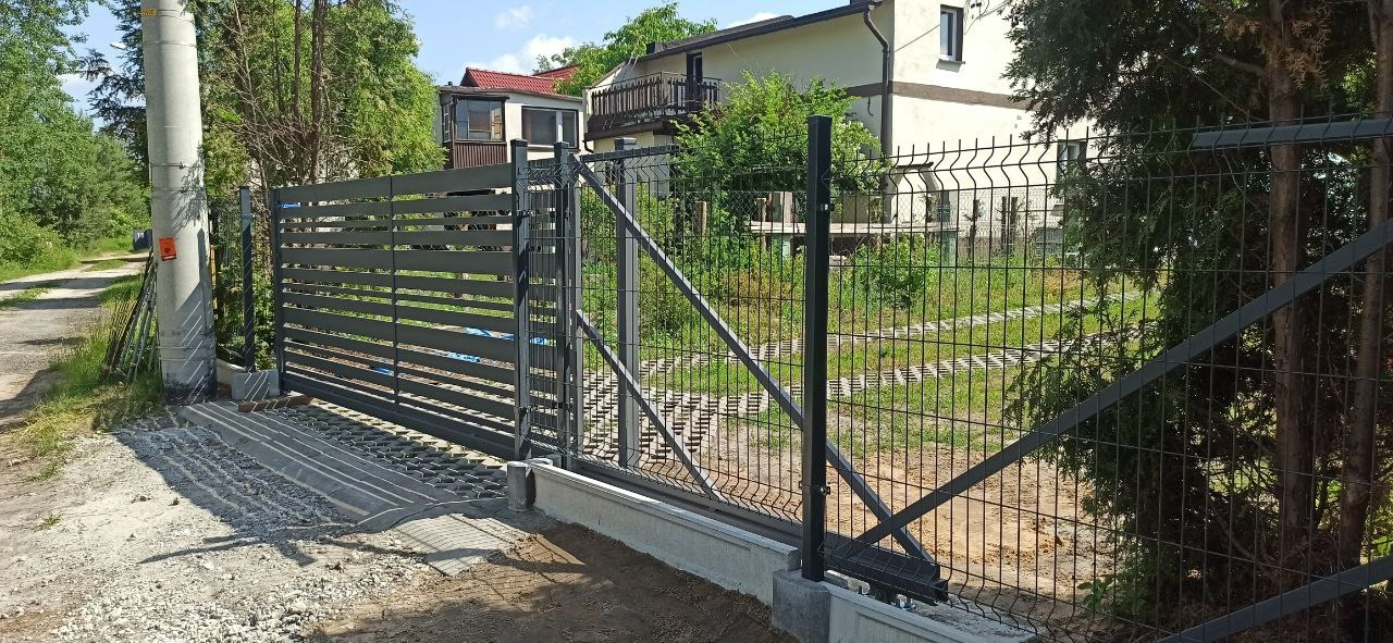 Nowoczesna przesuwna brama z poziomych profili i panelowe ogrodzenie w kolorze grafitowym, widoczne na tle posesji z domem jednorodzinnym i zielenią.
