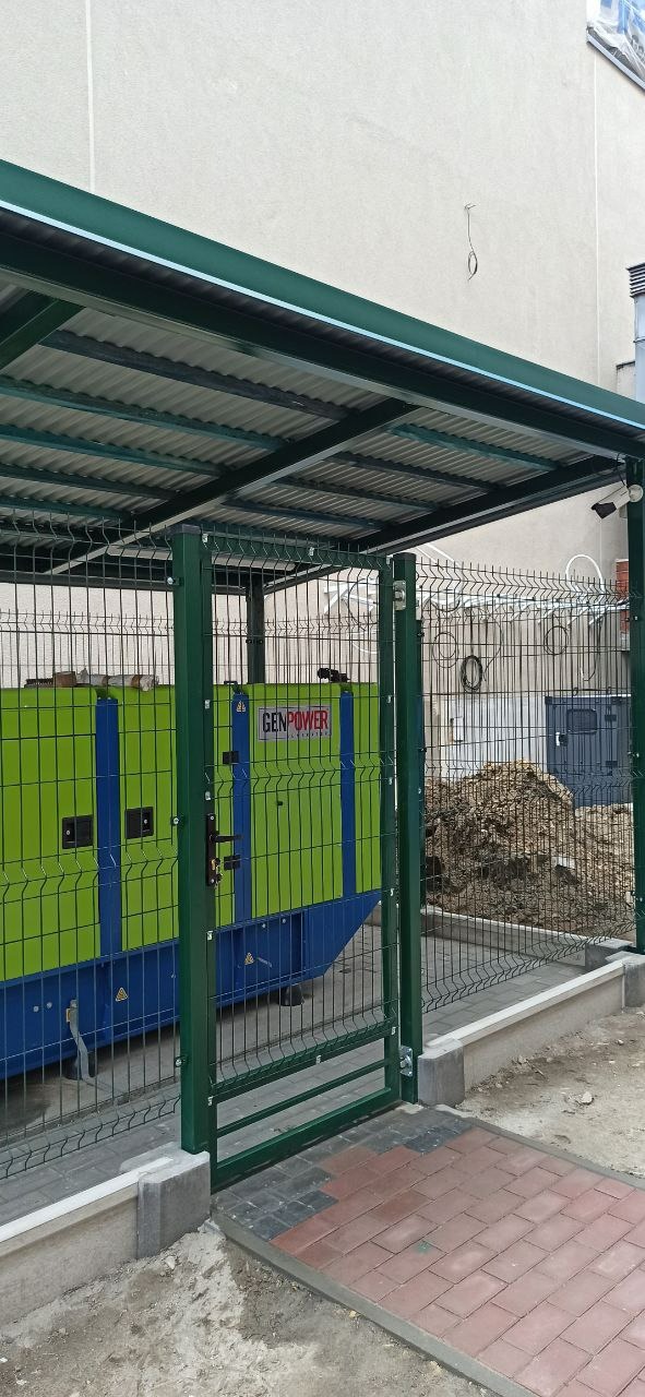 Zielony agregat prądotwórczy marki GenPower, umieszczony za ogrodzeniem z siatki, pod zadaszeniem z blachy falistej, z widoczną kamerą monitoringu.