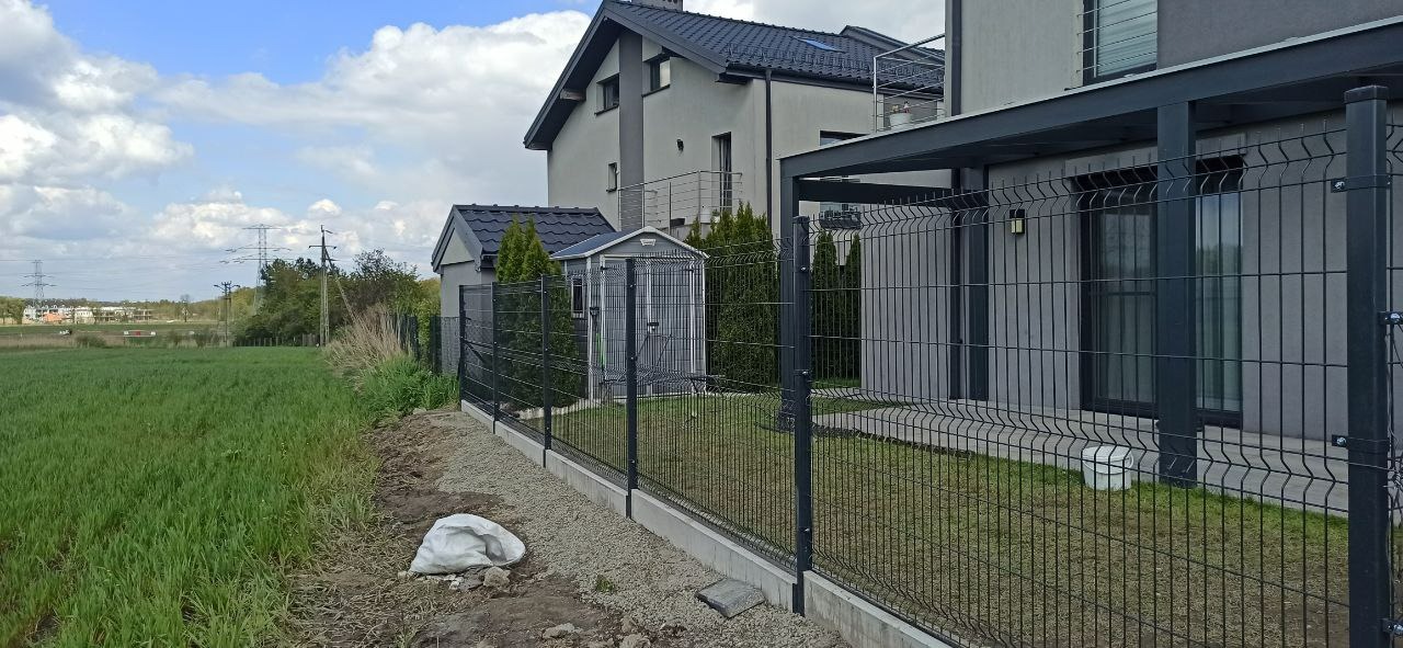 Czarny panelowy system ogrodzeniowy z podmurówką betonową, oddzielający posesję z domem w nowoczesnym stylu od pola uprawnego, z workiem gruzu przy fundamencie.