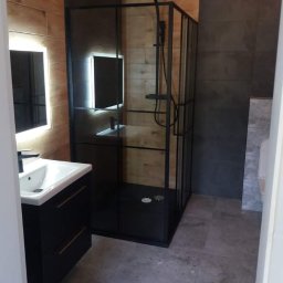 Retmix Poznań usługi budowlano - wykończeniowe - Nowoczesna łazienka z kabiną prysznicową typu walk-in z czarnymi profilami, umywalką nablatową na szafce w kolorze czarnym, podświetlanym lustrem i płytkami imitującymi drewno na ścianie.