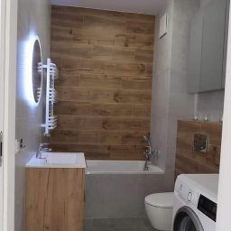 Retmix Poznań usługi budowlano - wykończeniowe - Nowoczesna łazienka z drewnopodobnymi płytkami na ścianie, okrągłym lustrem z podświetleniem LED, umywalką z szafką w kolorze drewna, białą wanną i pralką.