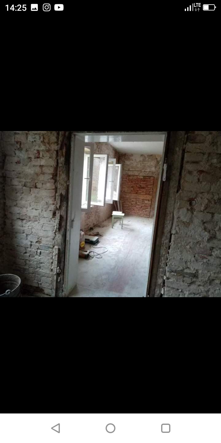 Wnętrze w trakcie remontu z odsłoniętą cegłą, widoczne okna, drabina i narzędzia na podłodze, surowe ściany z widocznymi śladami prac.