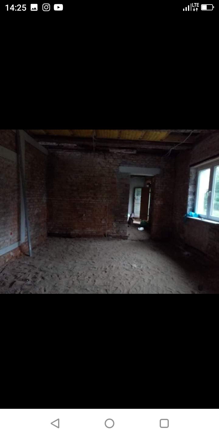 Wnętrze pomieszczenia w stanie surowym: ceglane ściany z widocznymi przewodami, okno z widokiem na zieleń, podłoga pokryta piaskiem, przygotowywane do dalszych prac.