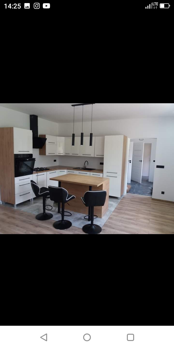 Nowoczesna kuchnia z białymi szafkami z drewnianymi akcentami, czarnym okapem i trzema czarnymi hokerami przy drewnianej wyspie; widoczne także drzwi do kolejnego pomieszczenia.