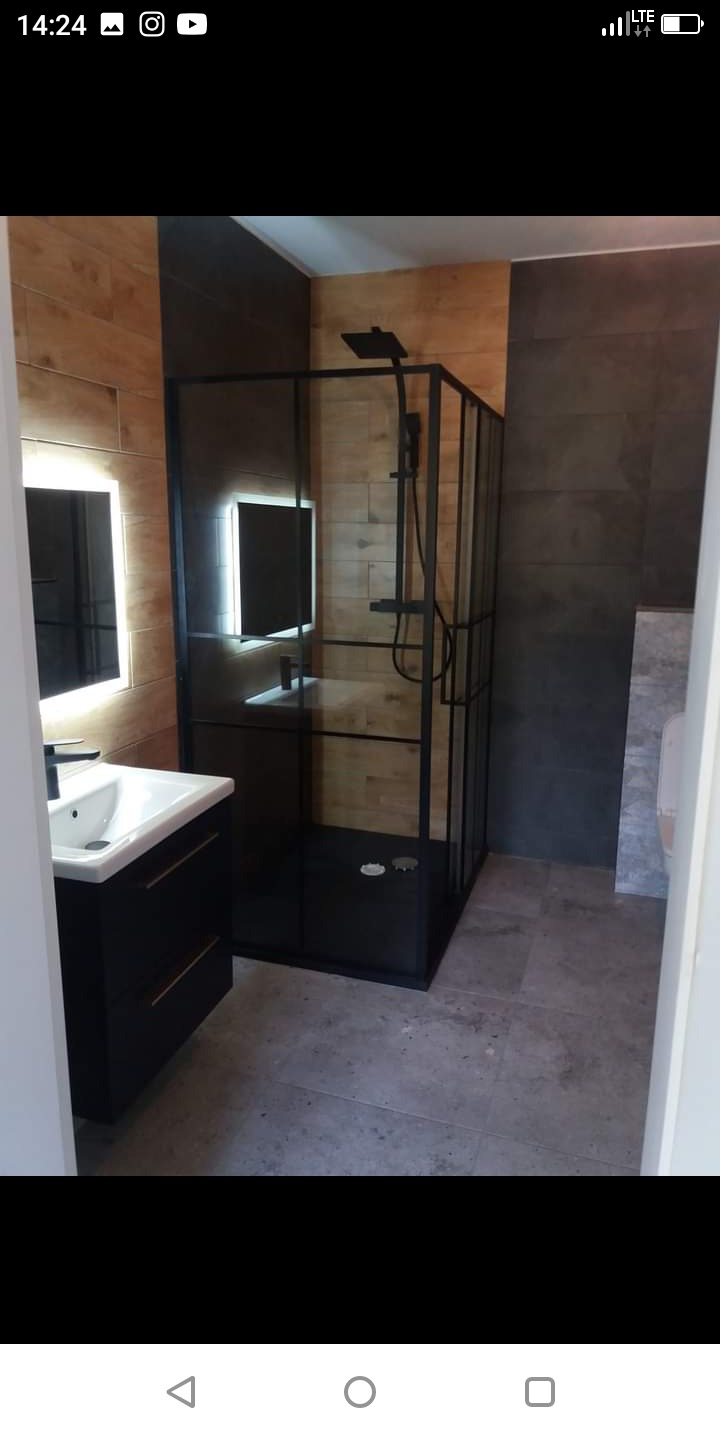 Nowoczesna łazienka z kabiną prysznicową typu walk-in z czarnymi profilami, umywalką nablatową na szafce w kolorze czarnym, podświetlanym lustrem i płytkami imitującymi drewno na ścianie.