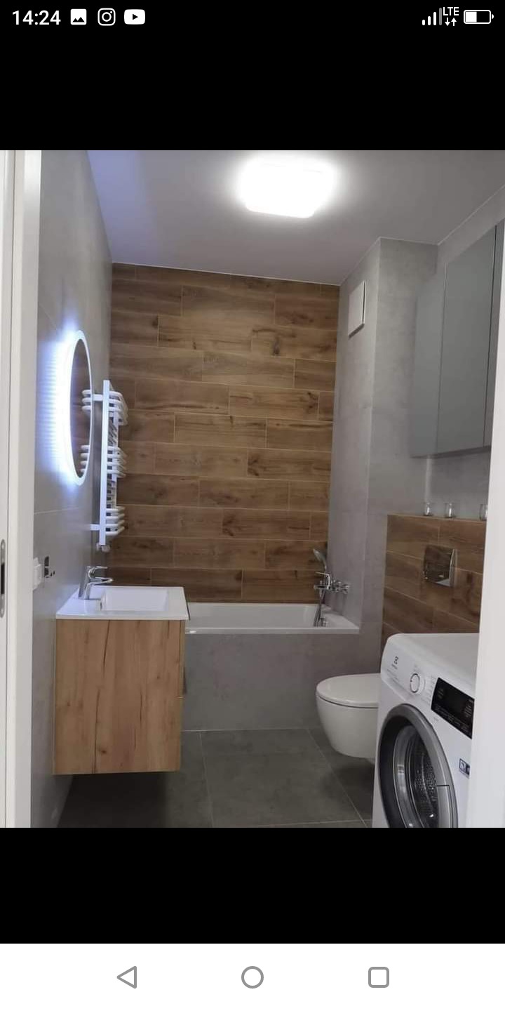 Nowoczesna łazienka z drewnopodobnymi płytkami na ścianie, okrągłym lustrem z podświetleniem LED, umywalką z szafką w kolorze drewna, białą wanną i pralką.