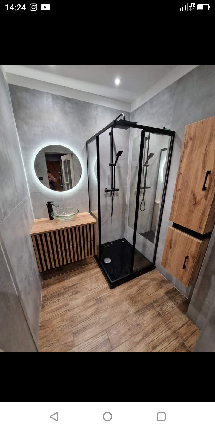 Nowoczesna łazienka z czarną kabiną prysznicową typu walk-in, drewnianą szafką pod umywalkę z okrągłym lustrem LED i drewnianymi szafkami wiszącymi.