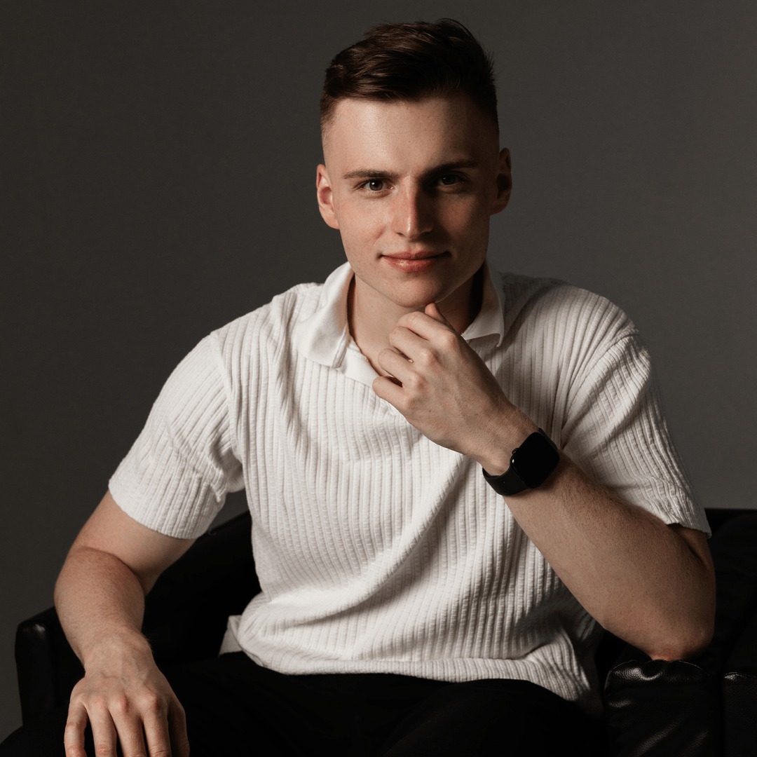 Młody mężczyzna w białej koszulce polo siedzi na czarnym skórzanym fotelu, zamyślony, dotykając brody, z czarnym smartwatchem na lewym nadgarstku, na szarym tle.