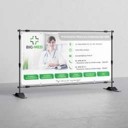 PawJan Design - Baner reklamowy przychodni medycznej BIG-MED z uśmiechniętą lekarką, danymi kontaktowymi i oferowanymi usługami, umieszczony na czarnym stojaku na tle szarej ściany.