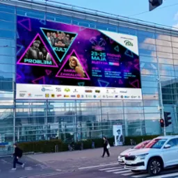 Wielkoformatowy baner reklamowy Juwenaliów Poznań 2024 z wizerunkami artystów i informacjami o wydarzeniu, umieszczony na fasadzie budynku, widoczny z perspektywy ulicy z przechodniami i samochodami.