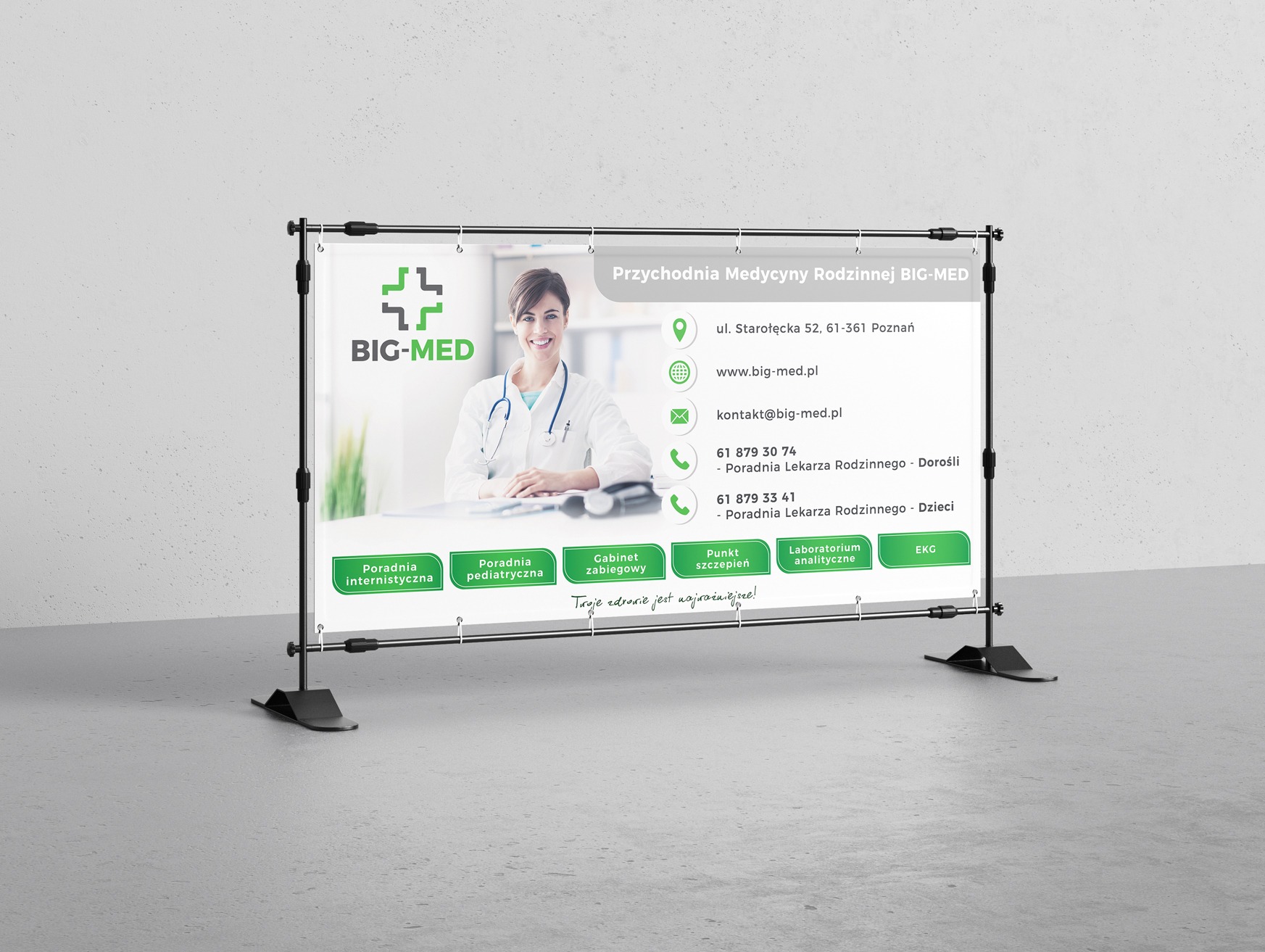 Baner reklamowy przychodni medycznej BIG-MED z uśmiechniętą lekarką, danymi kontaktowymi i oferowanymi usługami, umieszczony na czarnym stojaku na tle szarej ściany.