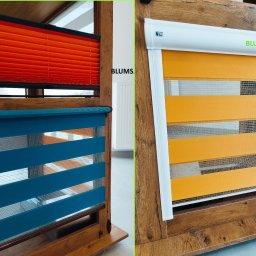 Blums - Ekspozycja dwóch typów osłon okiennych: pomarańczowe plisy i niebiesko-żółta roleta dzień noc, obie zamontowane w drewnianych ramach, z widocznym logo 'Blums'.
