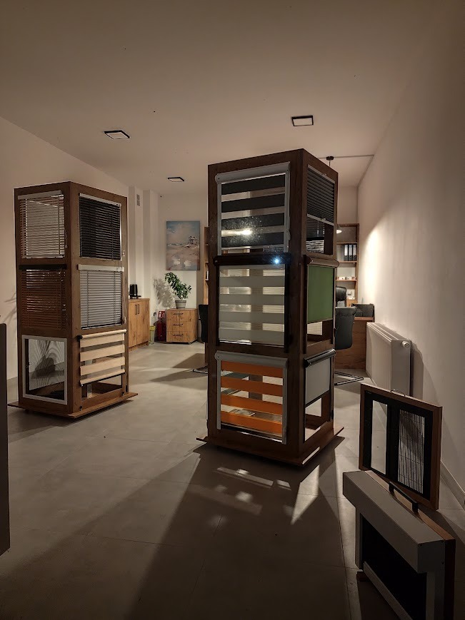 Ekspozycja w salonie z drewnianymi stojakami prezentującymi różne rodzaje osłon okiennych: poziome żaluzje drewniane, rolety dzień i noc, plisy oraz moskitiera w ramce.